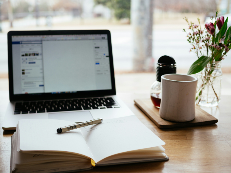 Picture of laptop on desk with an open book and pen in front of it